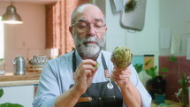 Senior food blogger removing artichoke leaves and speaking on camera, giving online culinary class or filming cooking vlog in the kitchen. Medium shot
