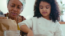 Little African-American girl mixing dough ingredients in glass bowl, baking together with cheerful senior grandmother during the day at home
