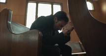 Young, emotional, anxious, and stressed man with long hair and black suit sitting in old church in worship and praying.