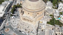 A far view, aerial 4k drone, footage tilt revealing the Mosta Rotunda Dome, a Roman Catholic church, and the surrounding city blocks of Malta near it.