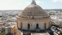 Closeup aerial 4k drone footage of the Mosta Rotunda Dome, a Roman Catholic church, ascending and moving forward to reveal the surrounding city of Malta.