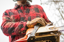 Man in red plaid jacket with sawdust with gloved hands holding chainsaw cutting wood 
