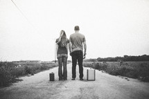 Couple standing in the middle of a dirt road with suitcases and a Bible.