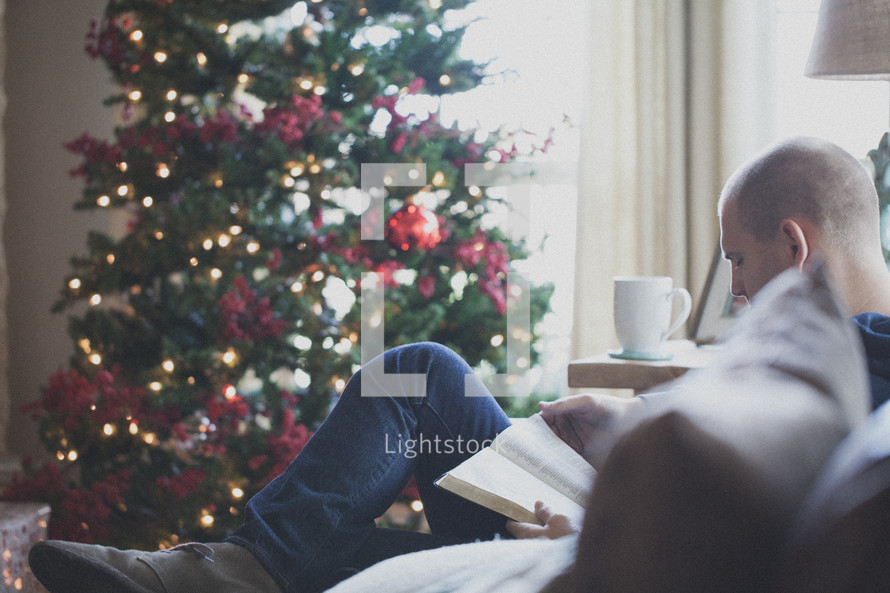 man reading a Bible on a couch in front of a Christmas tree 