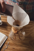 Coffee being poured into a chemex for brewing, next to a Bible and cross pendant.