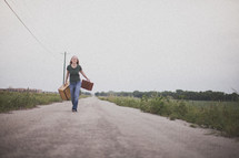Woman walking down the middle of a dirt road holding suitcases.