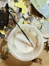artist's brush resting in glue bowl with torn and cut collage papers on work table