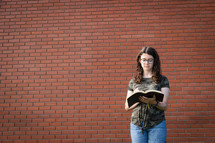 a teen girl reading a Bible standing in front of a brick wall 