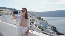 Woman taking phone selfie On travel in Oia, Santorini using smartphone. Female tourist sightseeing enjoying summer vacation visiting landmark destination in Greece, Europe
