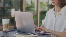 Cropped shot of woman typing on laptop while working at table in outdoor coffeeshop with notebook, pen, earphones and latte on it
