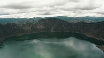 Aerial backwards shot of natural crater lake surrounded by volcano rocks during cloudy day	