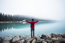 man standing on the shore of Lake Louise with hands raised 