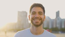 Handsome man looking at camera. Closeup positive guy smiling at camera. Portrait of male model posing on camera in urban sunset background. Cheerful man face with positive emotion

