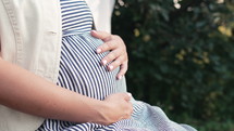 Pregnant woman sitting in summer home garden. Future mom caressing belly bump, green nature background. Expectant mother in summer dress, happy motherhood. High quality 4k footage