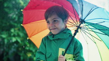 Happy preschool child boy under umbrella during rain. Smiling kid in raincoat enjoys autumn weather. Colorful parasol, raindrops, carefree childhood.