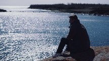 a man sitting on a rock looking out at the ocean 