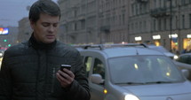 Man using his smart phone on a city sidewalk with traffic behind him.