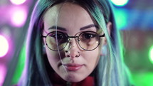 Millennial enigmatic pretty girl with unusual dyed hairstyle near glowing neon wall at night. Mysterious hipster teenager in glasses