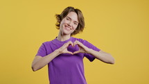 Smiling pretty woman showing sign of shape heart, sending love. Charming lady on yellow background. Women health, volunteering, charity donation, gratitude symbol, flirting concept. High quality 4k