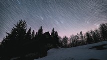 Time lapse of stars trails over wooden hut in forest.
