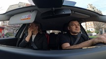 Real-life driving from a wide-angle camera captures a Caucasian family of three in their car, cruising through a city. The woman attentively applies eyebrow pencil while the man smiles and engages in conversation with her