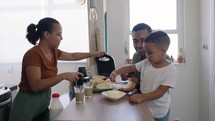 Family making breakfast while husband and little son eating and drinking juice at kitchen table