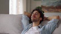 Happy relaxed young man rest lounge lean on couch enjoy peaceful mood, healthy lazy calm guy dreaming breathing fresh air sit on comfortable sofa in living room on stress free cozy day at home
