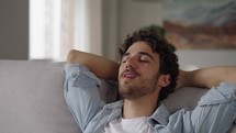 Happy lazy young man relax daydream meditate sit on comfortable sofa at home in living room, calm guy rest breathing fresh air having nap with eyes closed feel peace of mind, no stress free relief
