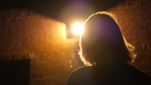 a woman standing outdoors walking towards a bright light at night 