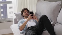Portrait of young man resting on sofa with a phone. Guy is laying on a couch uses mobile phone. Student studying online through phone at home.
