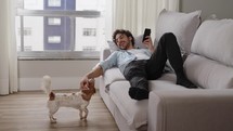 Caucasian man browses the web on a smartphone in the company of dog. A contented young boy uses phone while sitting on the living room couch smiling while stroking pet.
