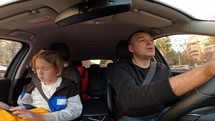 Real-life driving from a wide-angle camera - A Caucasian father in his 40s wearing a black t-shirt and his 12-year-old son are driving in a car, with the son engrossed in his phone and a yellow backpack on his lap, as they make a turn and drive around the city