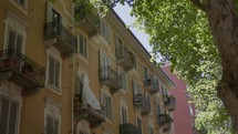 A white sheet hangs on the balcony with an iron railing, a yellow residential building, against the background of green foliage of a city street. Italy