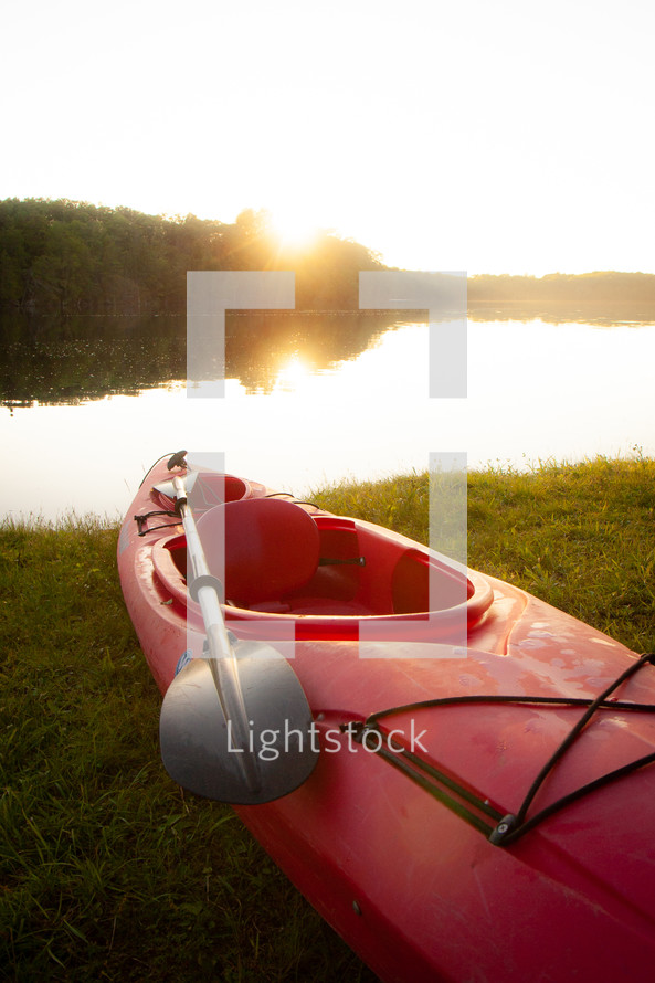 Red kayak with paddle oar on shore of lake at sunset