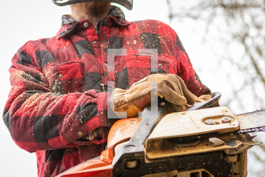 Man in red plaid jacket with sawdust with gloved hands holding chainsaw cutting wood 
