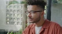 Medium close up shot of young multiracial man in glasses and casual outfit having talk with someone while sitting in cafe
