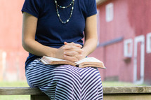 Young Christian woman sitting outside having quiet time with folded hands praying with open Bible in lap 