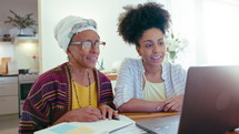 Young African-American woman sitting with senior mother at home, both smiling, waving and talking on video call on laptop. Medium shot
