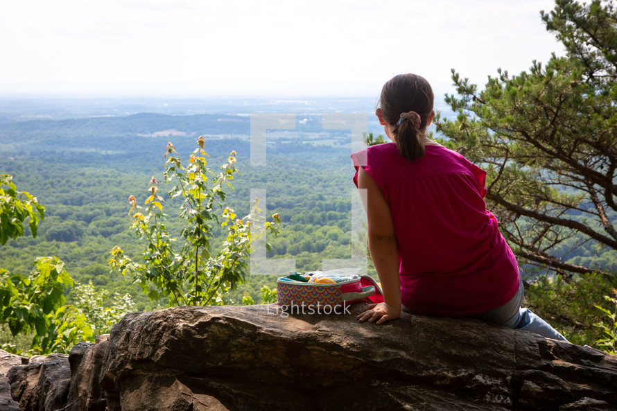 Young girl sitting and resting on cliff rock with picnic lunch looking out at forest overlook