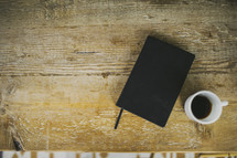 A black Bible and cup of coffee on a rustic wooden table.