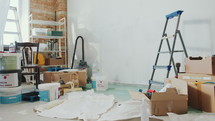 Interior of living room with cardboard boxes, paint buckets, ladder, tools and materials for home renovation. Zoom-out shot, no people
