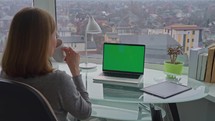 Woman sitting at workplace laptop with green display on the desk watching out of the window drinking coffee