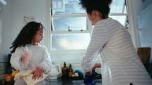 Cute little African-American girl sitting by the sink, drying dishes with towel, helping happy mother around the kitchen at home

