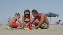 happy couple making sand castle at tropical beach with their son. Child with mom and dad outdoor at seaside playing together