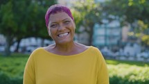 Happy senior stylish black woman smiling on camera in a public park. Pink hair
