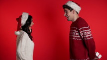 Young couple in Christmas Santa hats exchange gifts on red studio background