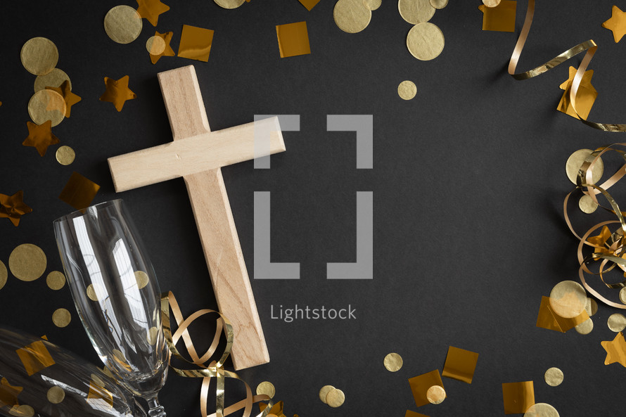 Wood cross and champagne flutes with gold streamers and confetti on a black background with copy space