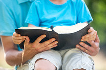 Father and young son on his lap reading the Bible together outside