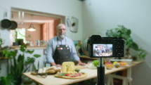 Senior male food blogger in apron standing by kitchen table with ingredients on it and talking, filming video recipe with digital camera on tripod. Zoom shot, rack focus
