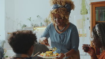 African American woman serving dessert on plates for relatives and talking to them during family dinner at home
